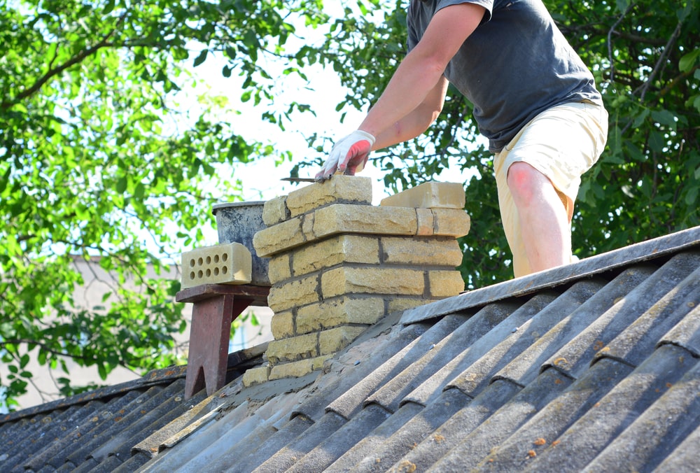 A person in a dark shirt, light shorts, and gloves works on brick chimney repair on a rooftop in Norfolk and Plymouth County, MA, surrounded by tools and green trees in the background.
