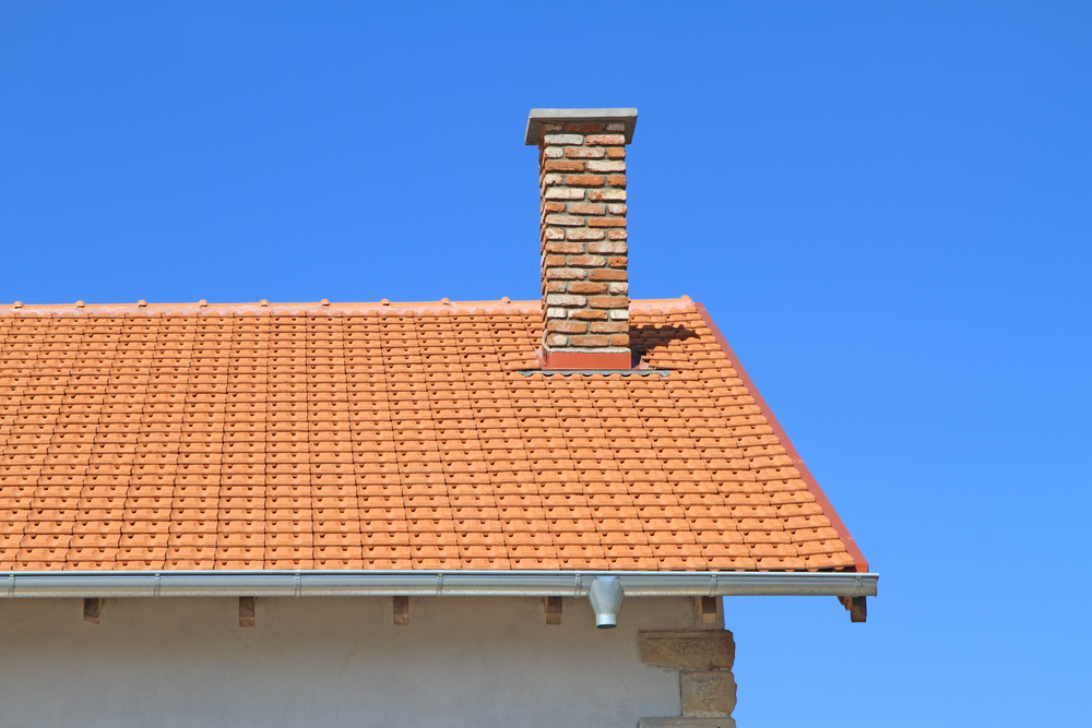 House roof with orange terracotta tiles and a tall red brick chimney, featuring a gutter and partial white exterior wall beneath a clear blue sky ideal for chimney restoration in Norfolk and Plymouth County, MA.