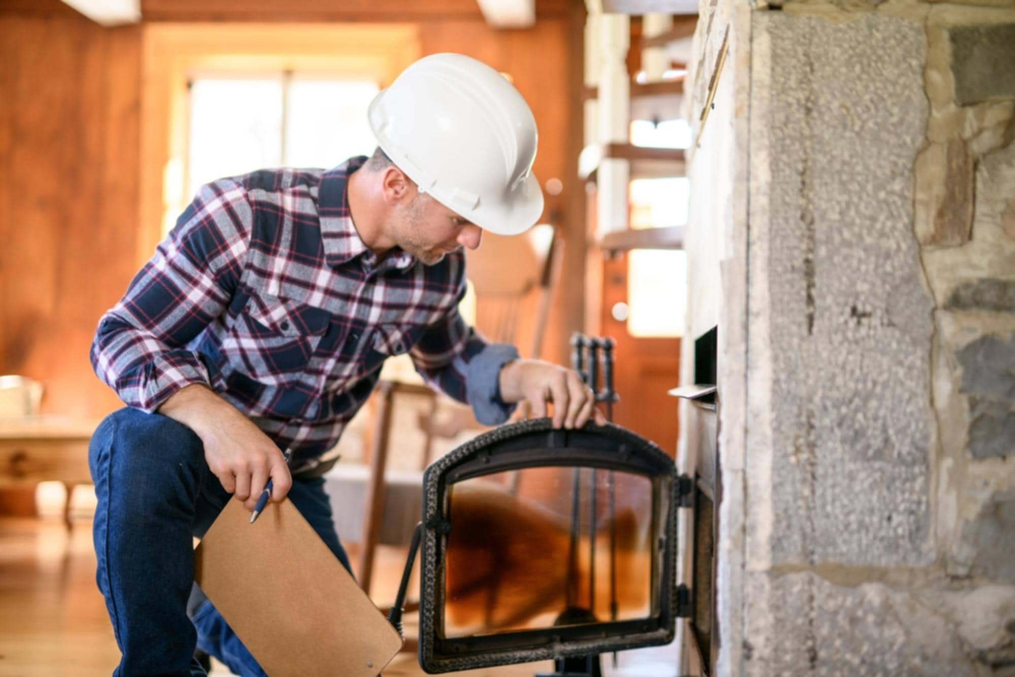 A man wearing a white hard hat and plaid shirt inspects a fireplace, holding a clipboard and opening the fireplace door inside a cozy, sunlit room with wooden furniture and stone walls.