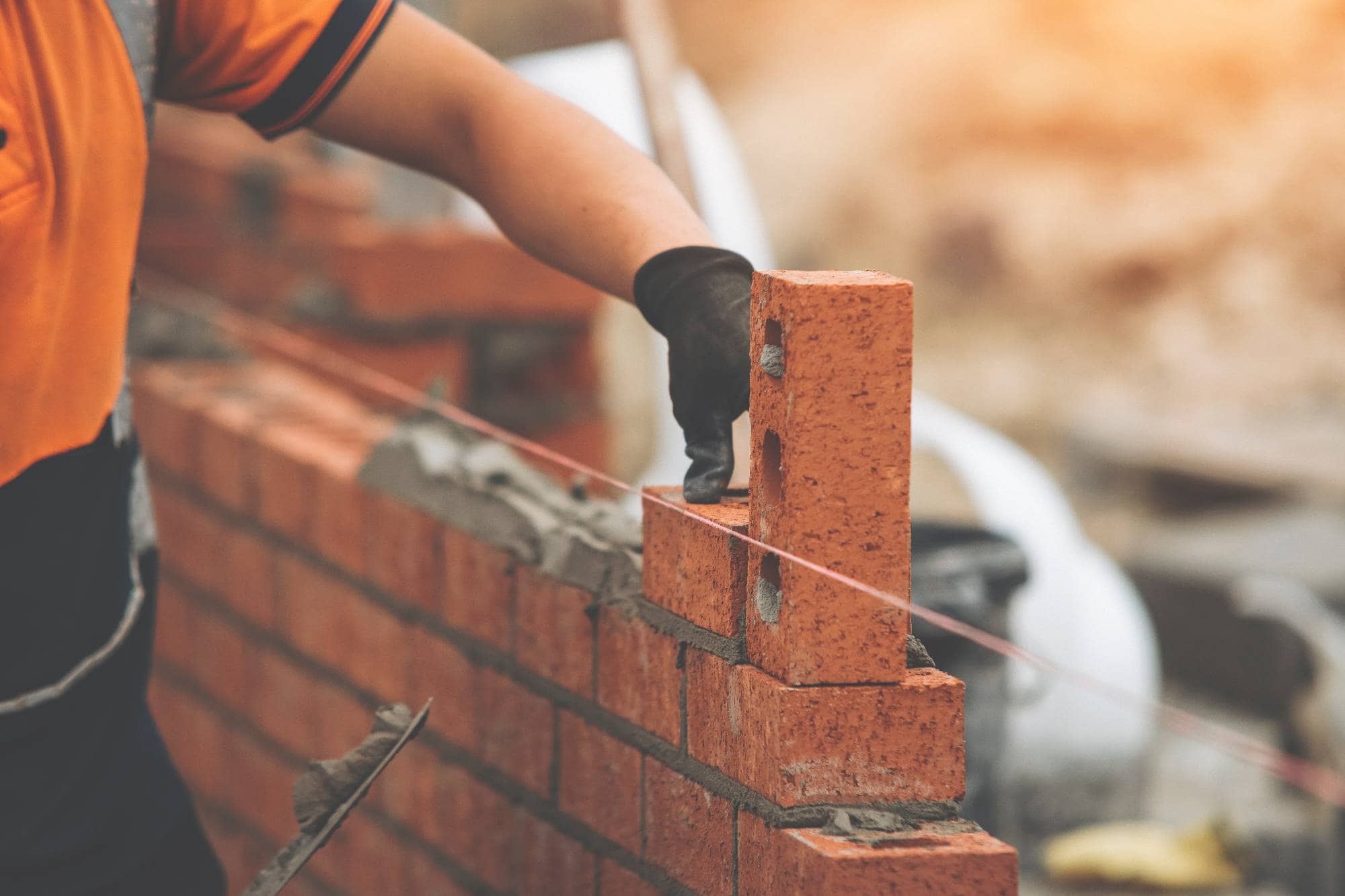 A construction worker wearing an orange shirt and black glove lays a brick on a brick wall, aligning it with a guide string.