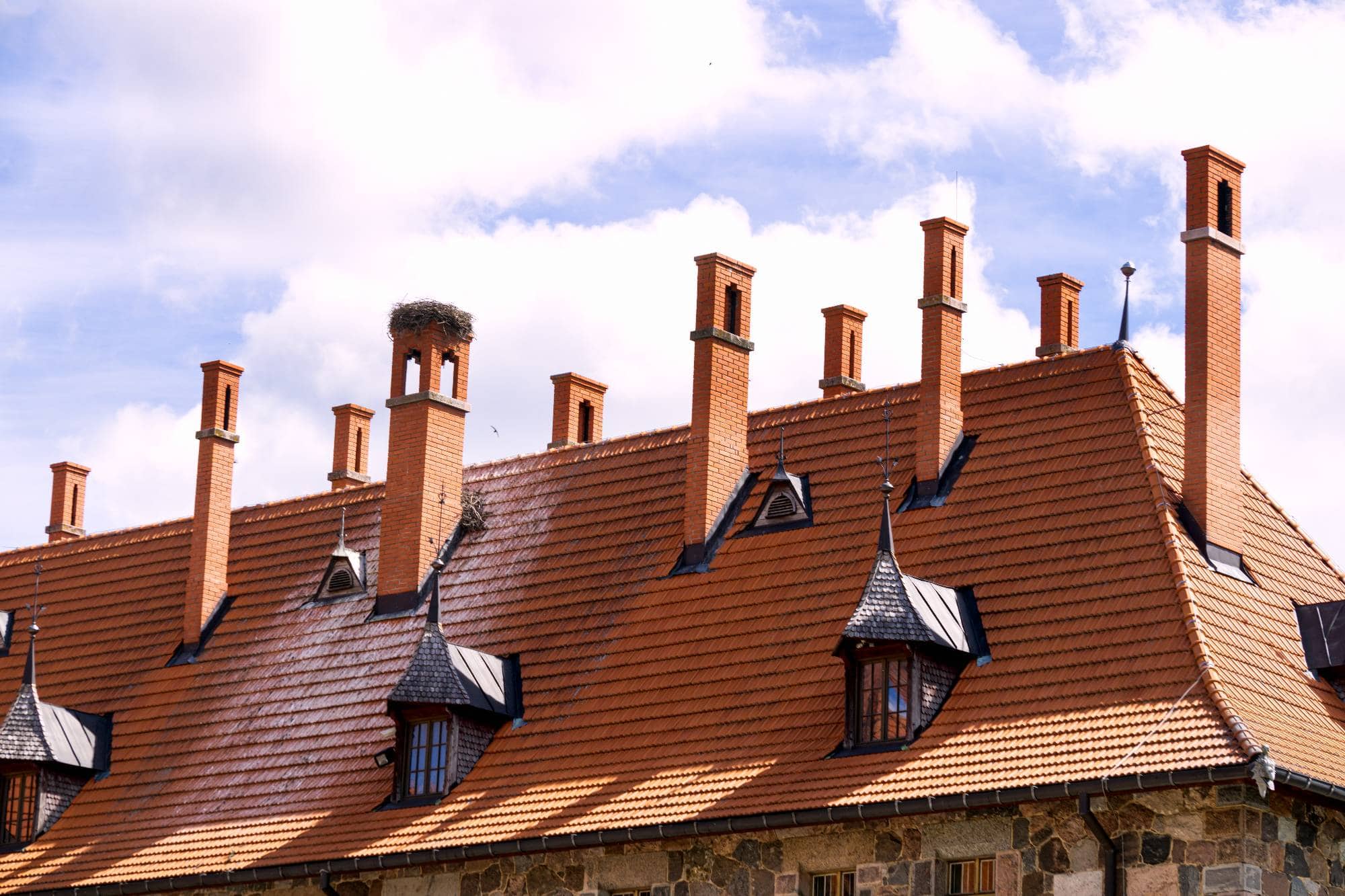 A building with a steep red-tiled roof, multiple tall brick chimneys, and small dormer windows. A large bird’s nest sits atop one chimney, set against a blue sky with scattered clouds.