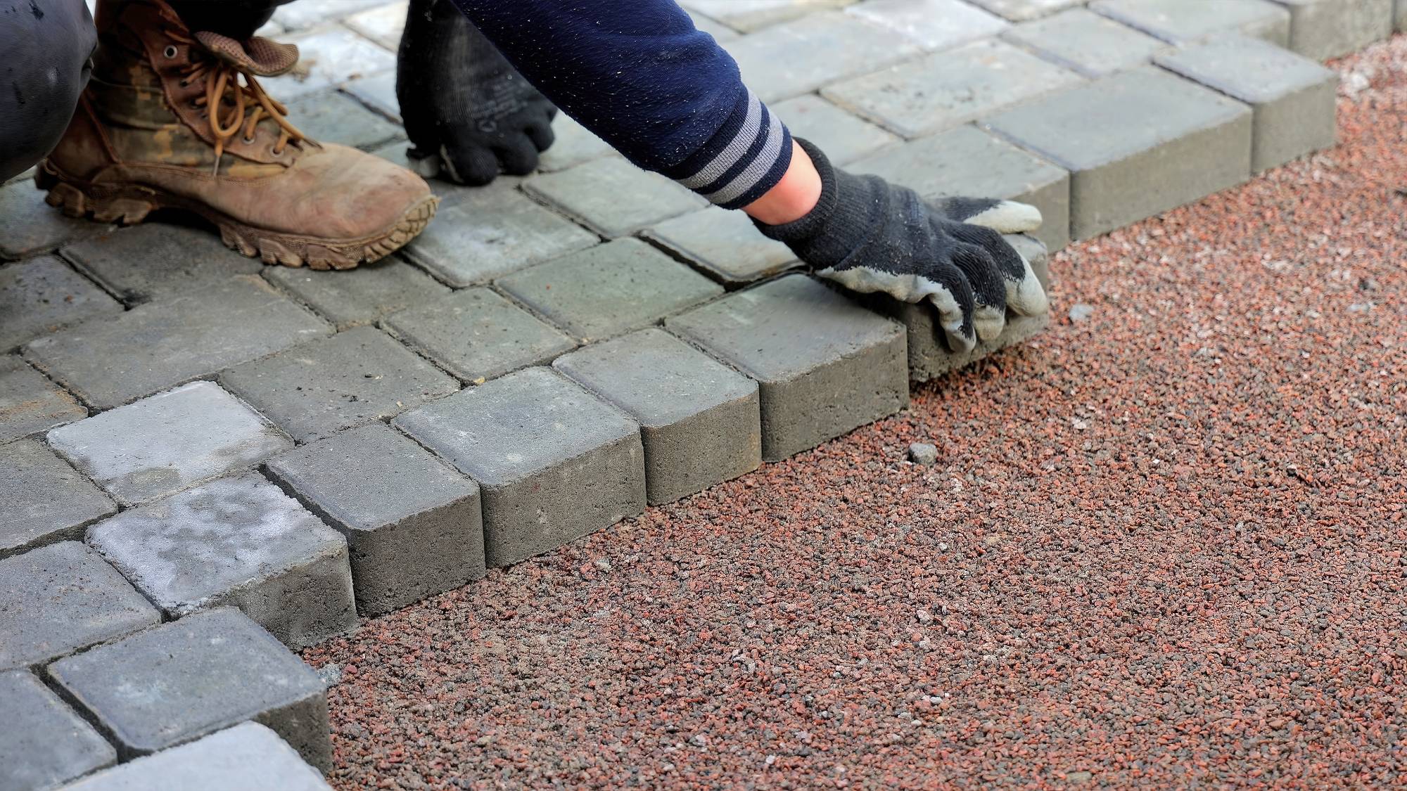 A person wearing gloves and work boots is placing gray paving stones onto a bed of reddish gravel, constructing a pathway or patio.