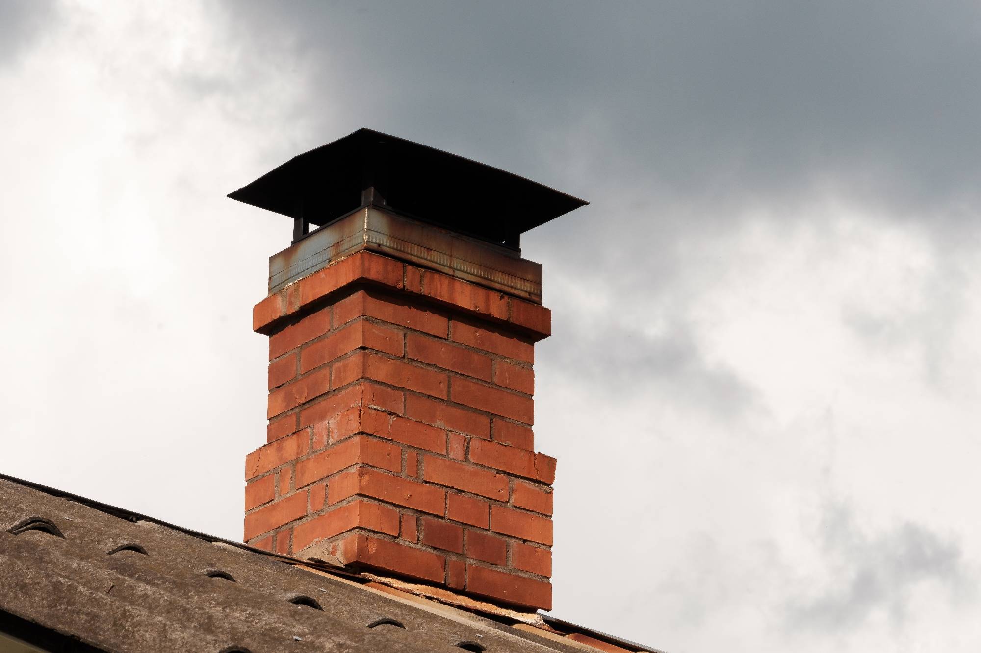 A red brick chimney with a black metal cap on a sloped roof, set against a cloudy sky.