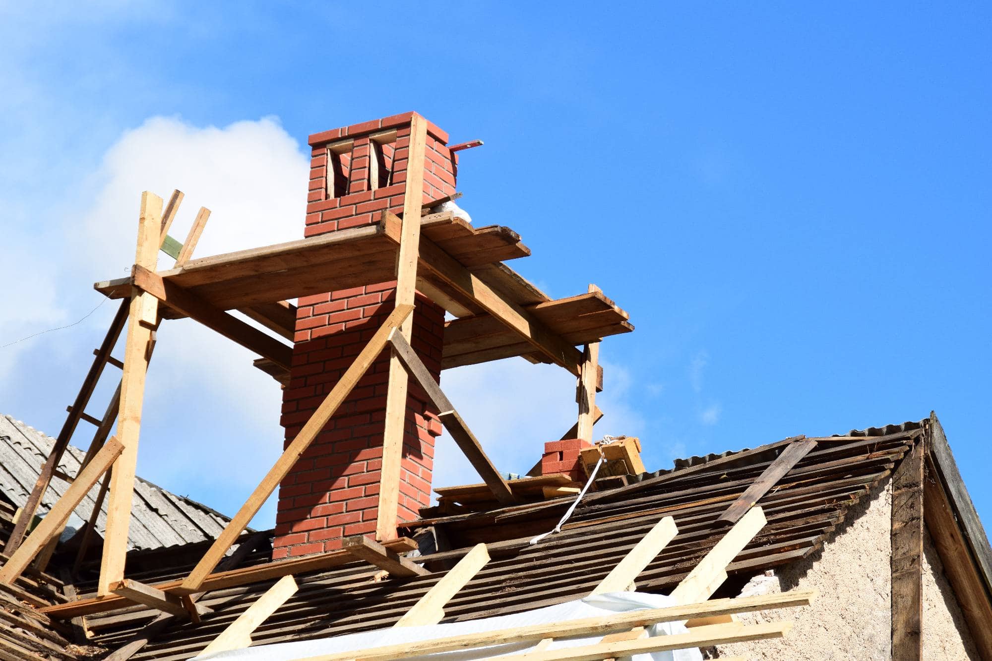 A brick chimney surrounded by wooden scaffolding is under construction on a house roof with missing tiles, against a bright blue sky with clouds.
