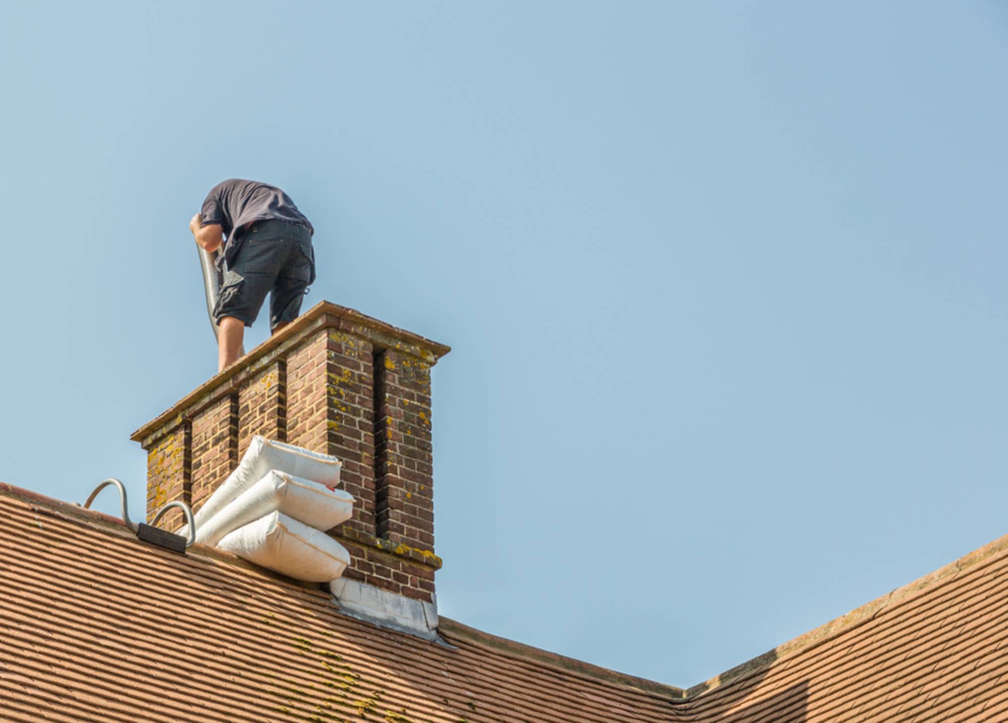 A person in black clothes stands on a roof, leaning over a brick chimney, with large white bags secured to the chimney, under a clear blue sky.