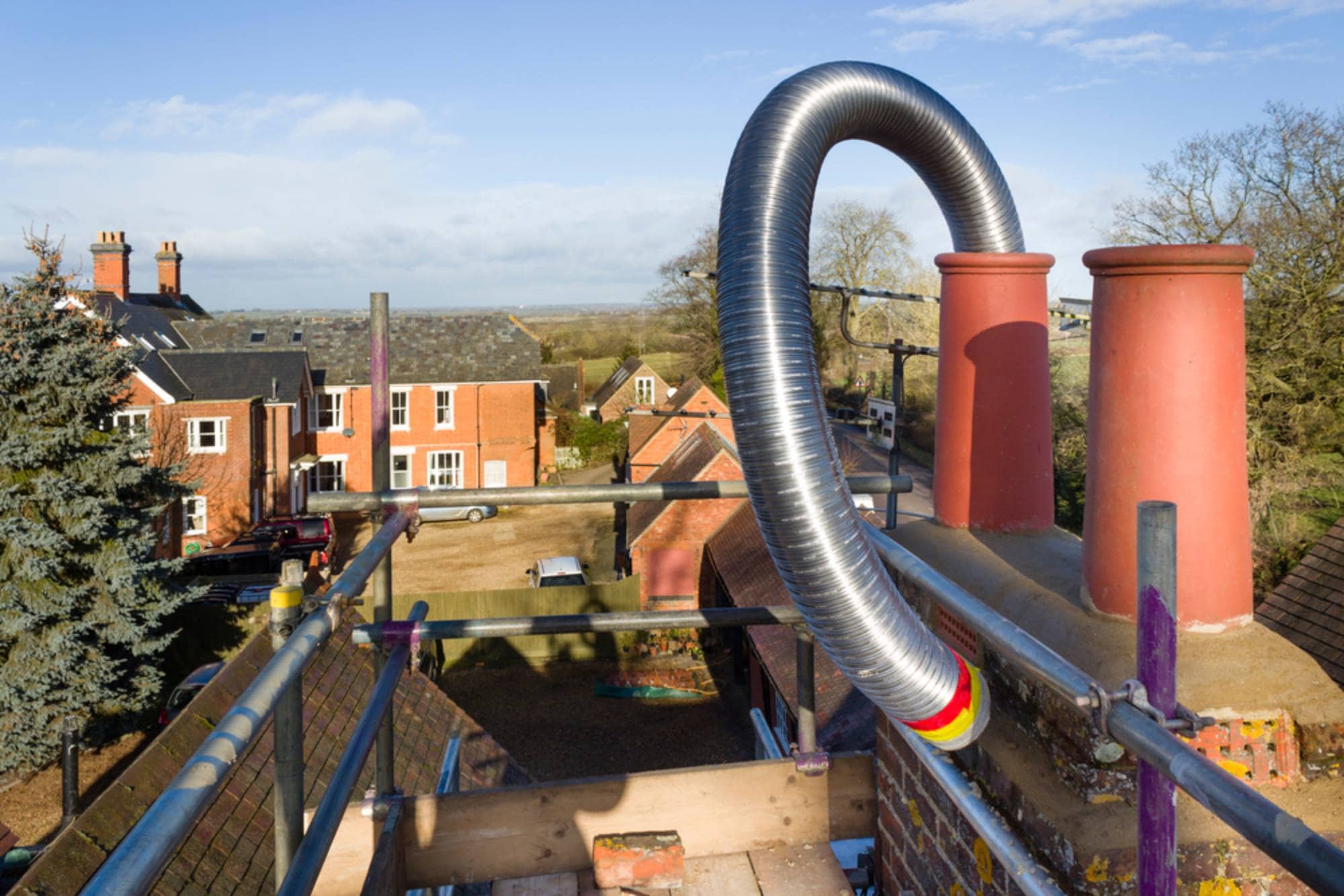 A flexible metal flue liner is being installed in a brick chimney on a rooftop, with red chimney pots and scaffolding. Houses, trees, and a clear sky are visible in the background.