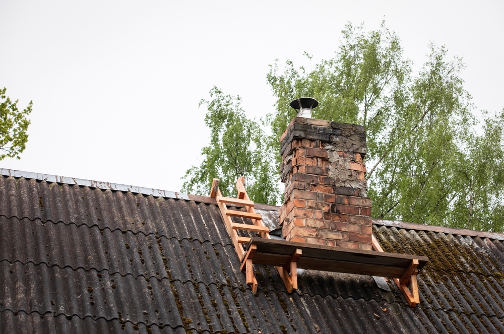 A brick chimney with a metal cap rises from a corrugated roof, hinting at recent chimney repair Norfolk and Plymouth County. A small wooden platform and short ladder sit nearby, while trees with green leaves frame the background.