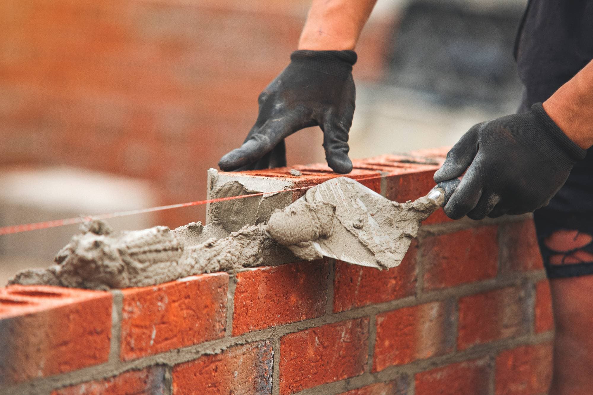 A person wearing black gloves uses a trowel to spread mortar on a brick wall under construction, with a guideline string visible for alignment.