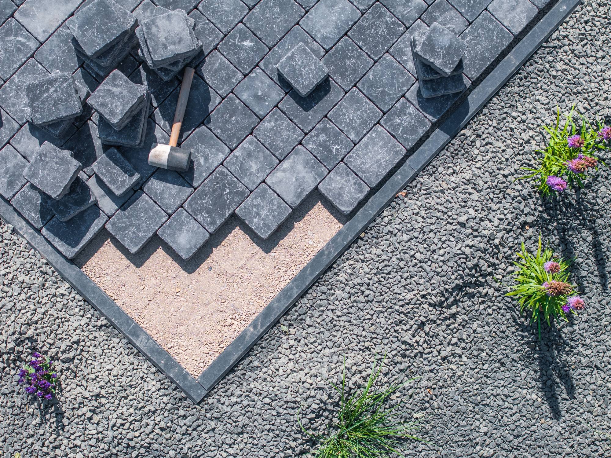 A patio is being built with dark stone pavers, some stacked and some placed. A rubber mallet rests on the pavers. Surrounding the patio are gravel and a few small flowering plants.