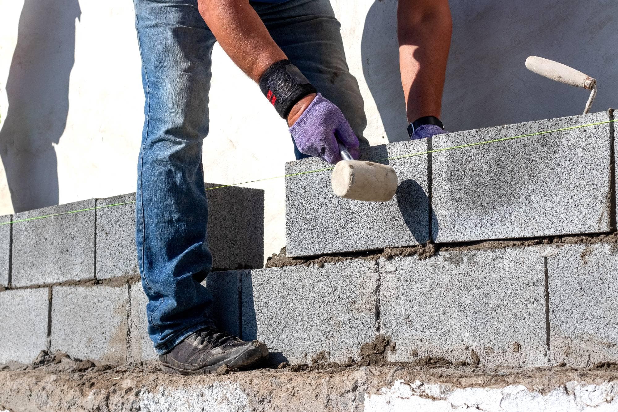 A construction worker wearing gloves and jeans uses a rubber mallet to set concrete blocks in a straight line, building a wall with mortar on a sunny day.