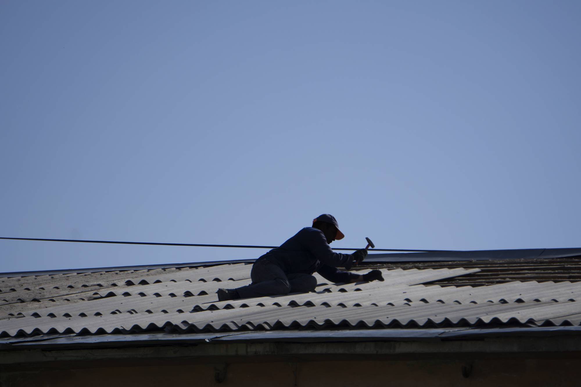 A person wearing dark clothing and a cap is kneeling on a corrugated metal roof, using a hammer to work, under a clear blue sky.