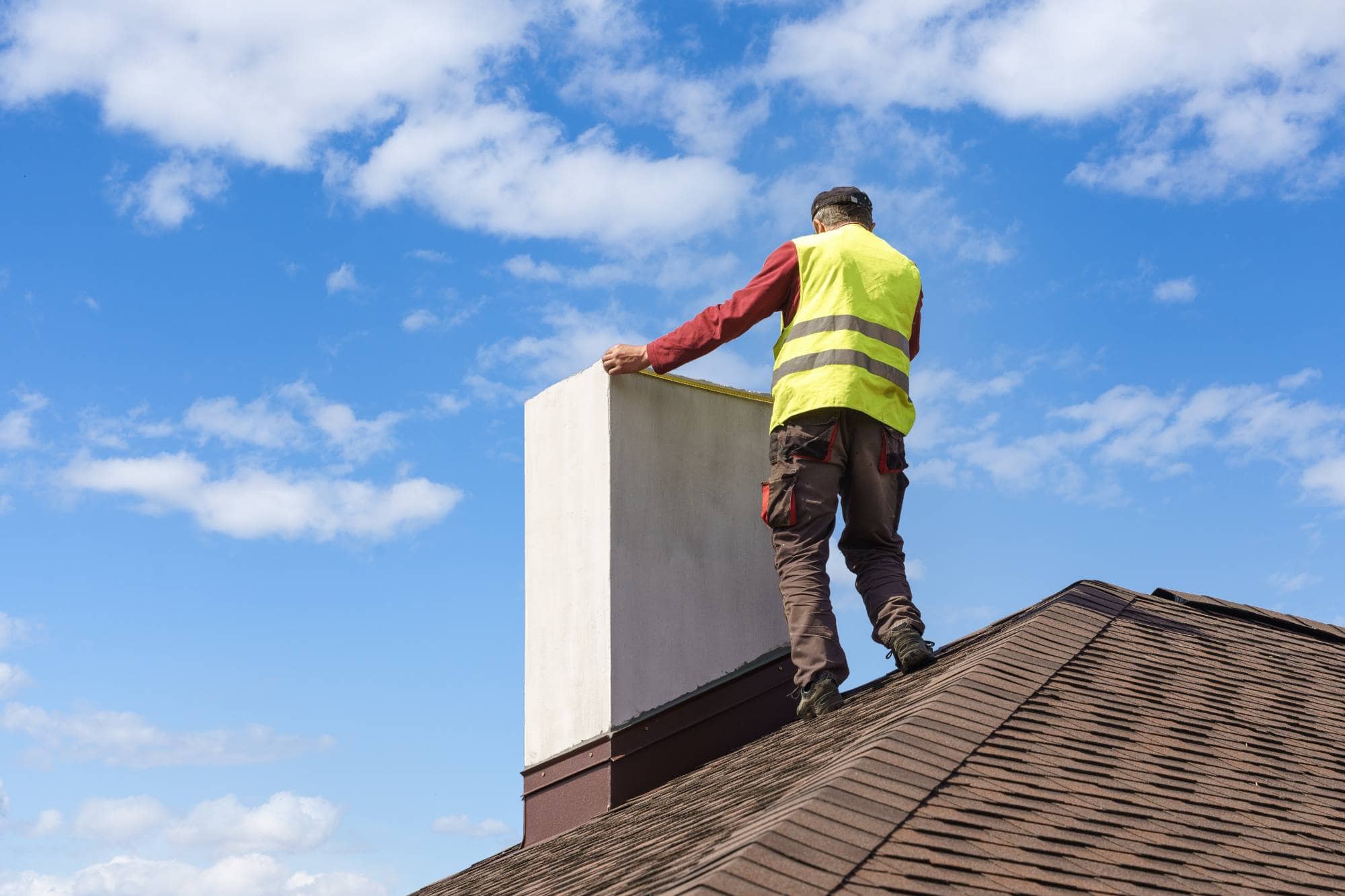 A worker in a yellow safety vest stands on a sloped roof, inspecting a white chimney against a blue sky with scattered clouds.