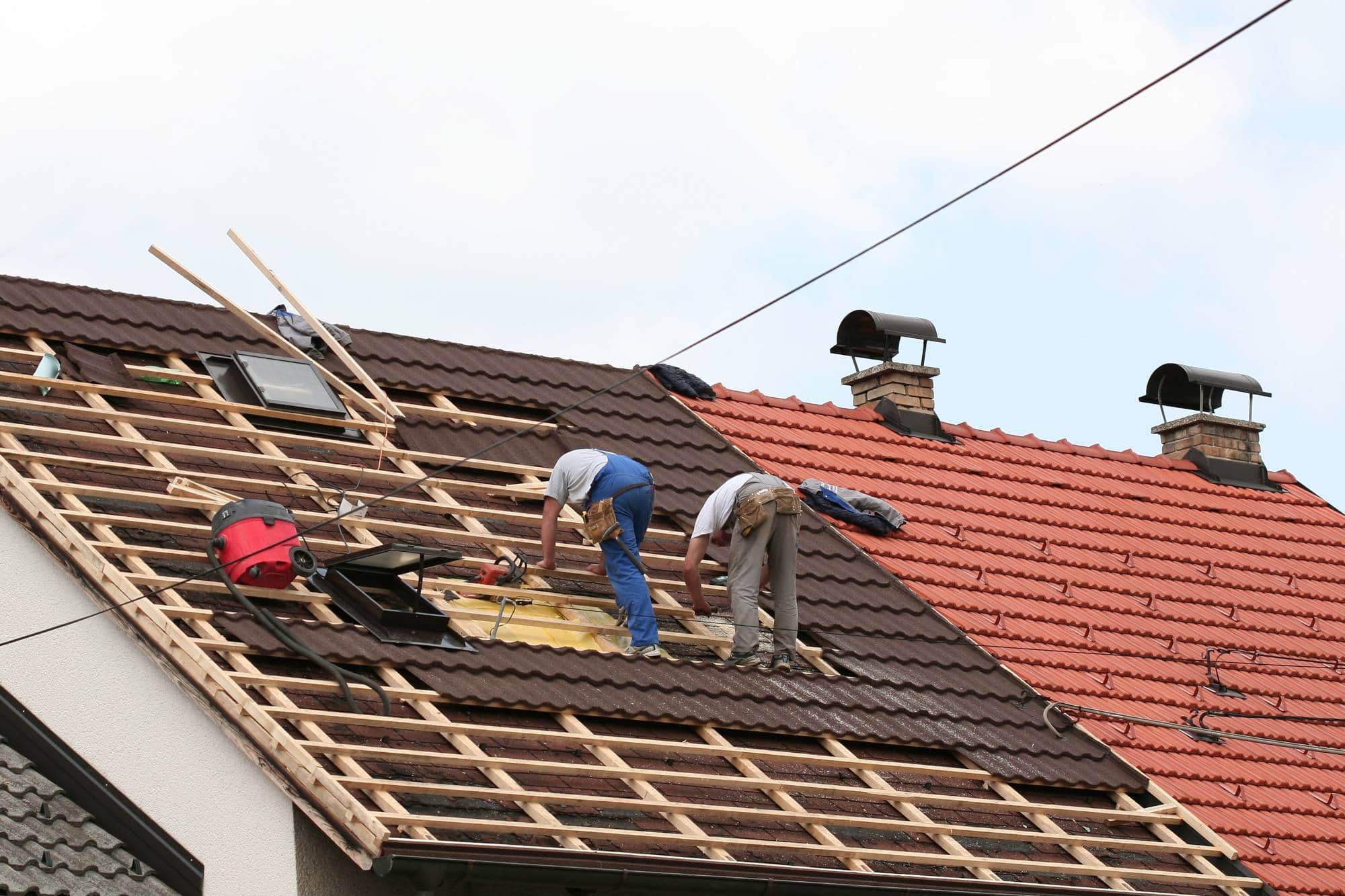 Two workers repair the roof of a house, surrounded by tools and wooden beams, with one section of tiles removed. Another roof with red tiles is visible beside them under a partly cloudy sky.