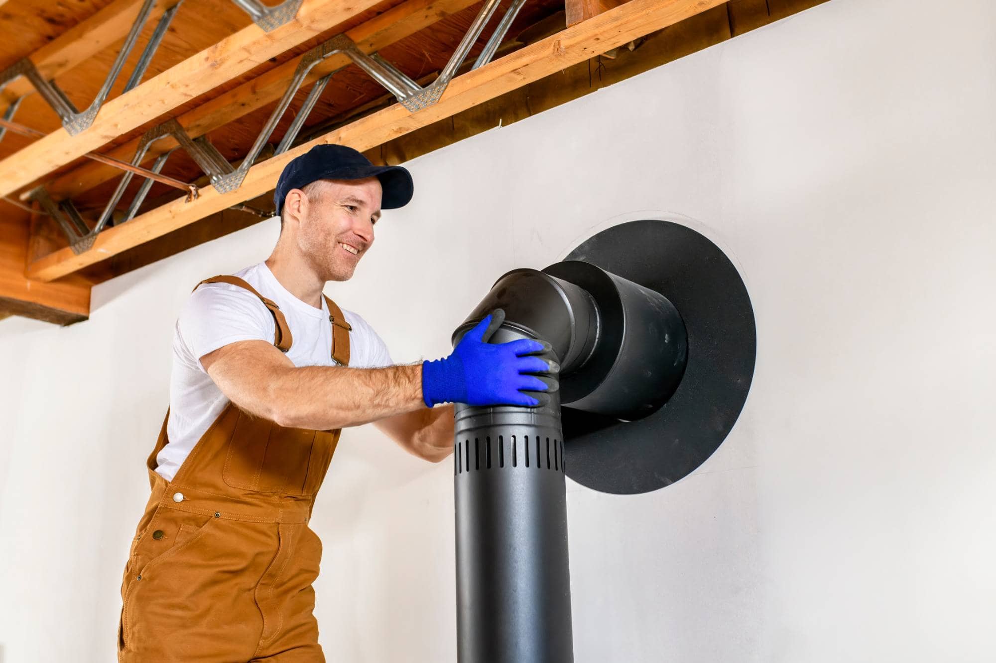 A smiling worker in brown overalls and blue gloves installs a black stove pipe into a wall opening in a room with exposed wooden beams on the ceiling.