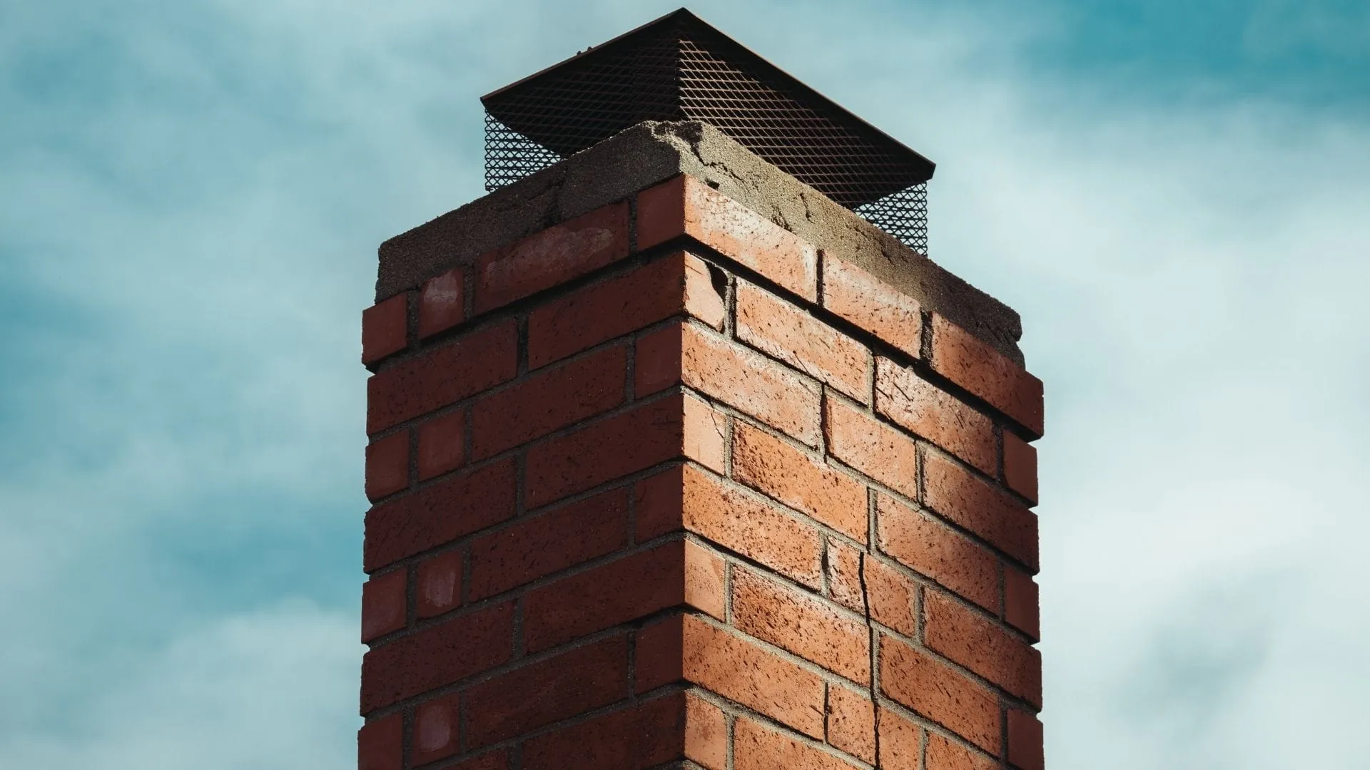 A close-up of a red brick chimney with a metal cap and mesh screen at the top, showing clearly defined mortar lines, set against a partly cloudy blue sky.