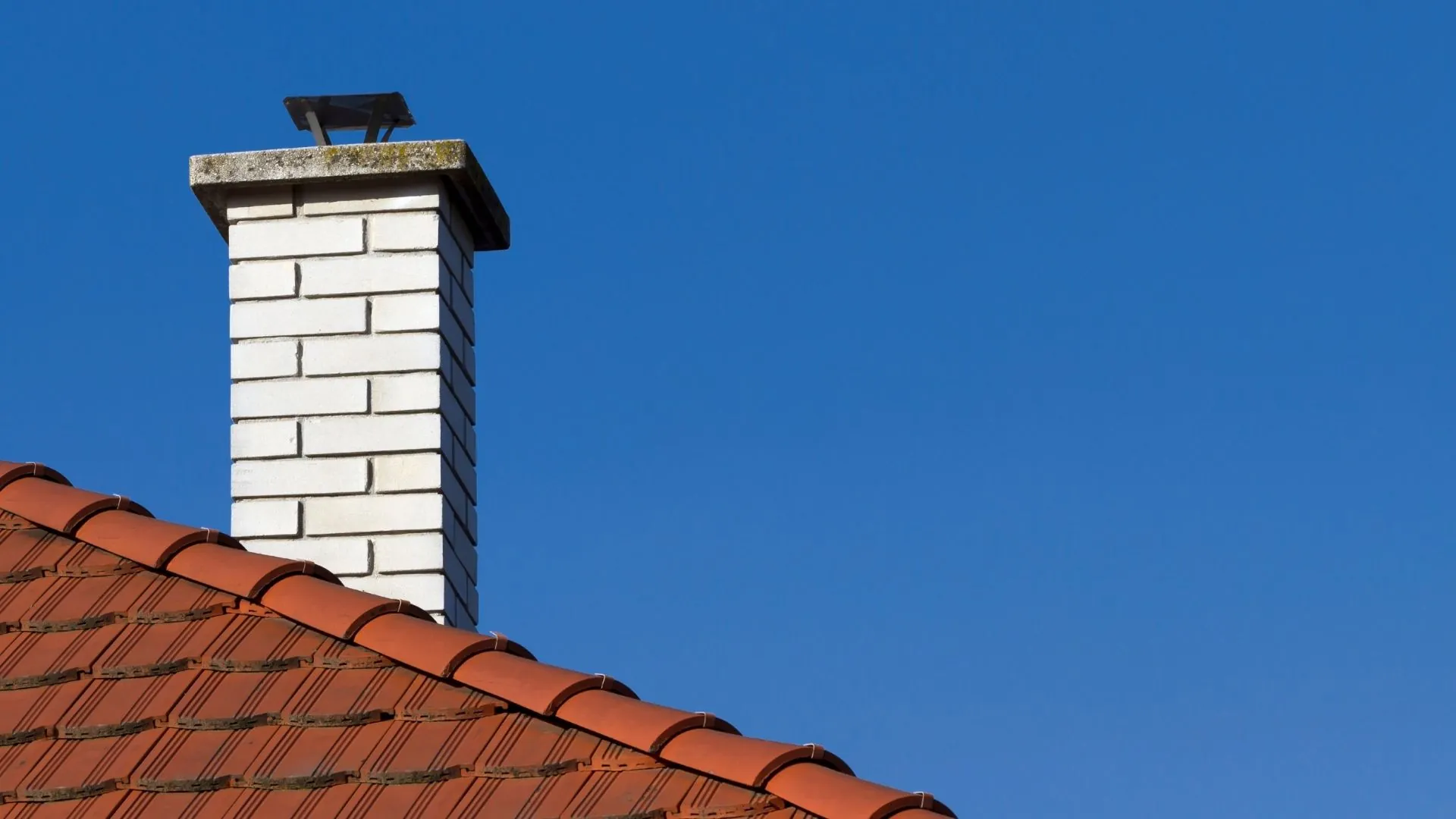 A white brick chimney with a metal cap rises above a red tiled roof against a clear blue sky.